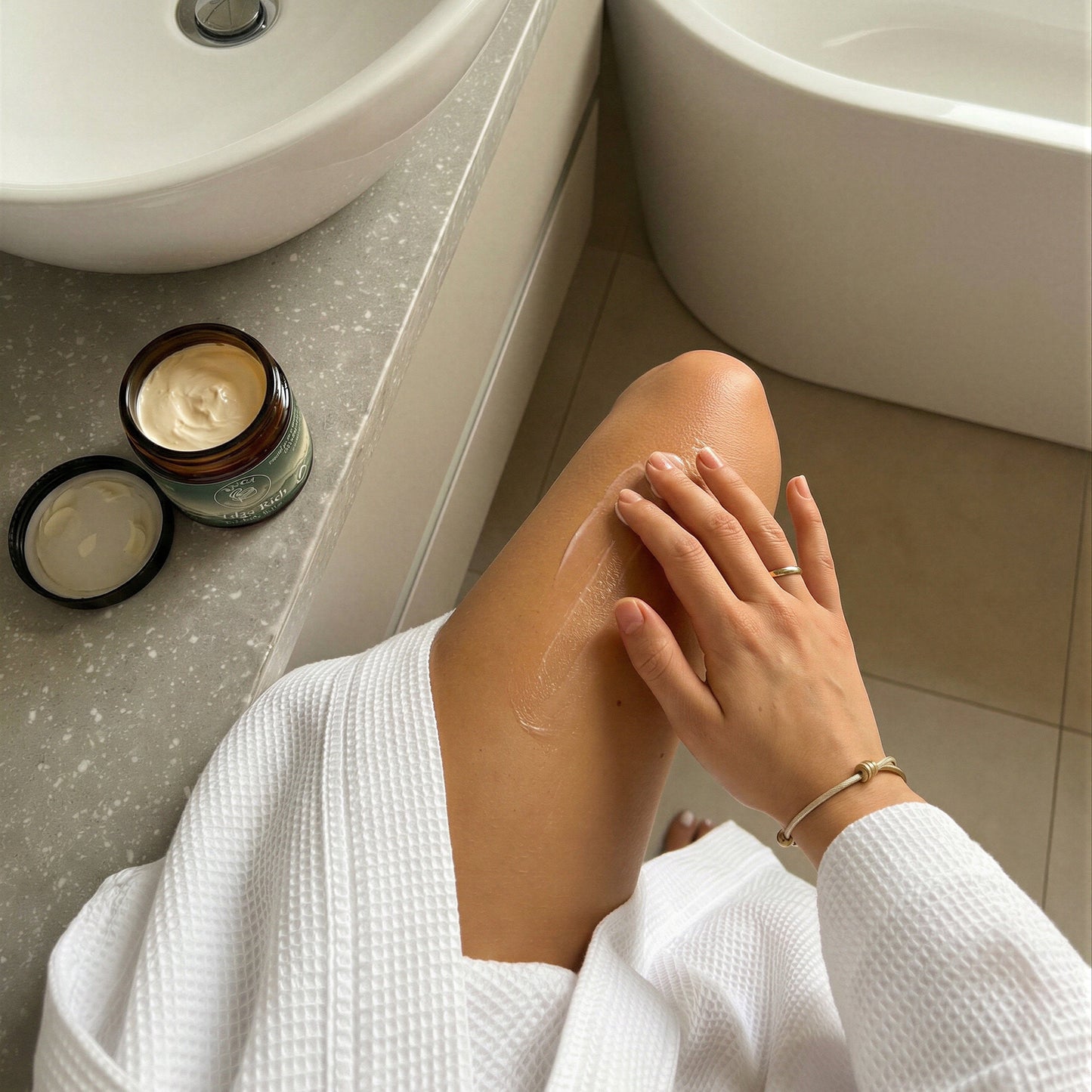 Person in a white robe applying Talga Rich cream to their leg beside an open jar on a bathroom counter.